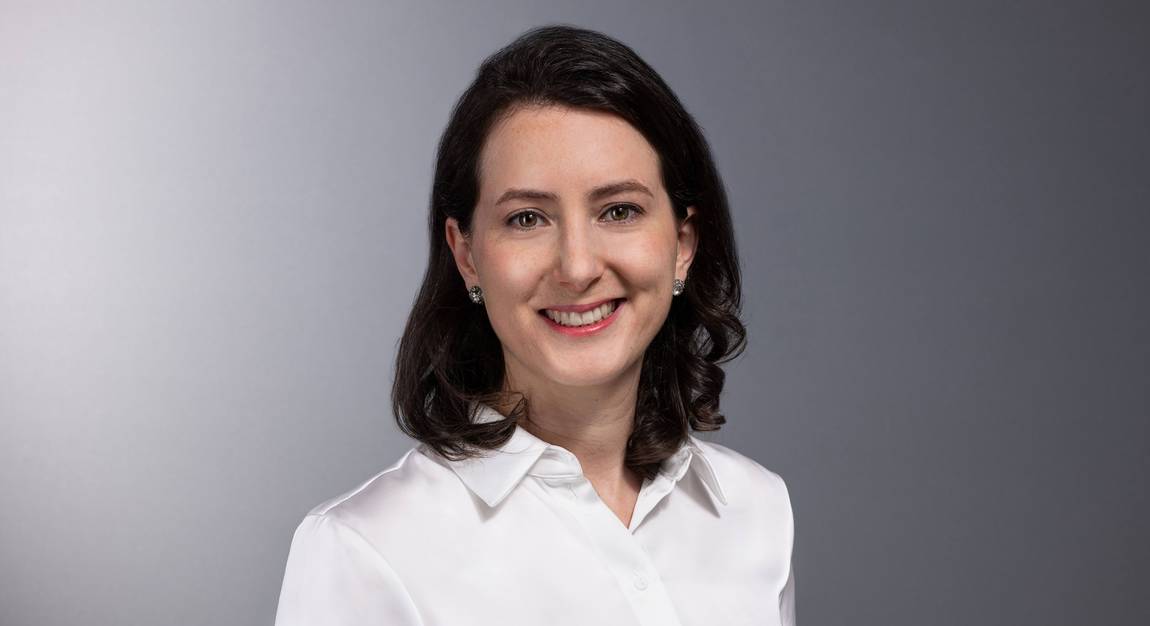 Eine Frau mit schulterlangen dunklen Haaren und weißem Kragenhemd lächelt in die Kamera vor einem schlichten grauen Hintergrund.